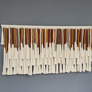May include: A wall hanging made of white yarn with brown, orange, and yellow accents. The yarn is woven into a series of tassels that hang down from a horizontal rod.