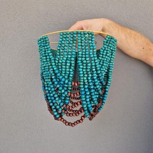 May include: A teal and brown beaded lampshade. The lampshade is held by a gold-colored metal frame. The beads are arranged in vertical strands, with teal beads at the top that transition to brown beads at the bottom. The background is a solid gray.
