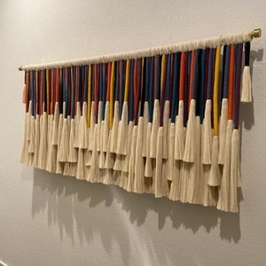 Puede incluir: Un tapiz de pared colorido hecho de hilo con borlas. El hilo es en tonos de rojo, naranja, amarillo, verde, azul, morado y blanco. Las borlas son todas blancas.