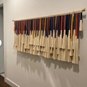 Puede incluir: Un tapiz de macram&eacute; de pared colorido con una variedad de hilos de lana de diferentes colores y borlas blancas. El tapiz de pared est&aacute; hecho de fibra natural y es una gran adici&oacute;n a cualquier decoraci&oacute;n del hogar.