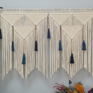 May include: A white macrame wall hanging with blue tassels and wooden beads. The hanging is made of cotton cord and has a geometric pattern.
