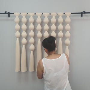 May include: A white macrame wall hanging with a series of round, puffy knots and long tassels. The hanging is attached to a black metal rod.