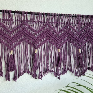 May include: A deep purple macrame wall hanging featuring a chevron pattern created with intricate knotted cord. The design includes vertical fringe strands, small light-colored wooden beads, and tassels suspended from a black horizontal rod.