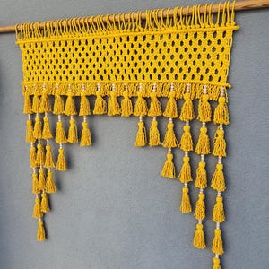 May include: A handmade yellow macrame wall hanging with a wooden dowel. The piece features a woven net design with multiple rows of yellow tassels, each adorned with small white beads. The wall decor is displayed against a gray wall.