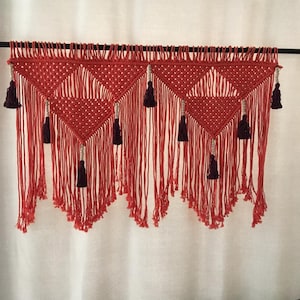 Puede incluir: Un tapiz de macramé rojo y blanco con borlas. El tapiz está hecho de cuerdas anudadas y tiene un patrón geométrico.