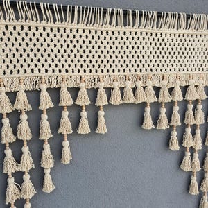 May include: Cream-colored macrame wall hanging with a woven design and fringe. The top section features a grid pattern, while the bottom has multiple tassels with wooden beads. The piece is suspended from a dark rod, creating a bohemian aesthetic.