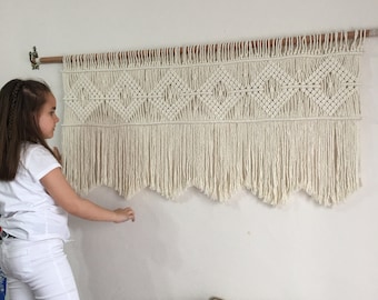 May include: A white macrame wall hanging with a geometric pattern and fringe. The hanging is mounted on a wooden dowel rod.