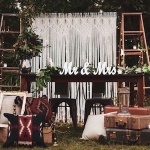 Könnte beinhalten: Eine rustikale Hochzeitszeremonie-Kulisse mit einem weißen Makramee-Vorhang, zwei Holzleitern und einem Holzschild mit der Aufschrift "Mr. & Mrs.". Die Kulisse ist mit Grünpflanzen und Lichterketten geschmückt.