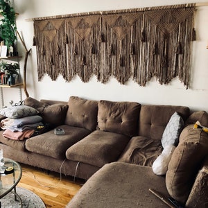May include: A brown sectional sofa with a worn fabric texture. The sofa is positioned in front of a wall with a large macrame wall hanging featuring a geometric pattern and tassels.