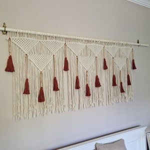 May include: A white macrame wall hanging with a geometric pattern and brown wooden beads. The hanging has red tassels along the bottom.