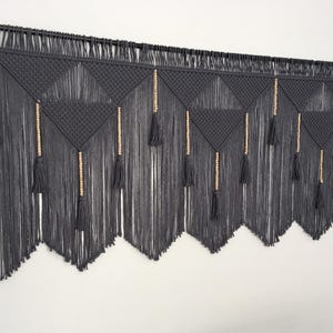 May include: A gray macrame wall hanging with wooden beads and tassels. The wall hanging is made of a thick cord and has a geometric pattern.
