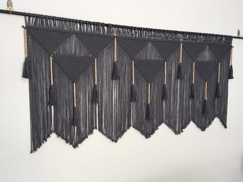 May include: A gray macrame wall hanging with wooden beads and tassels. The wall hanging is made of a thick cord and has a geometric pattern.
