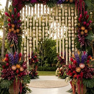 Könnte beinhalten: Ein Hochzeitsbogen mit einem Holzrahmen, der mit leuchtenden tropischen Blumen wie violetten Orchideen, orangefarbenen Proteen und rotem Ingwer geschmückt ist. Hängende Holzperlenstränge zieren die Mitte, flankiert von zwei großen Steinurnen mit üppigen Blumenarrangements.