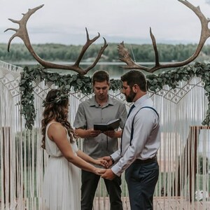 May include: A wedding ceremony taking place outdoors with a couple standing under a large antler arch decorated with greenery. The couple is holding hands and looking at each other. The ceremony is taking place in front of a lake.
