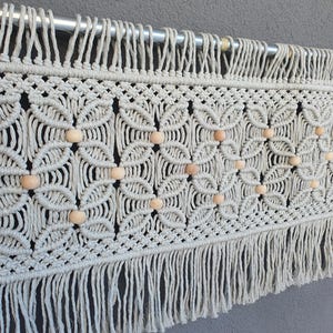 May include: A cream-colored macrame wall hanging with a geometric floral pattern and wooden beads. The piece is suspended from a silver metal rod and has a fringed bottom edge. This handcrafted textile adds a bohemian touch to any space.