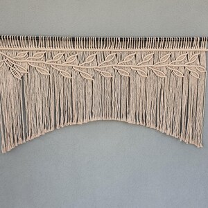 May include: A beige macrame wall hanging with a leaf design. The macrame is suspended from a black rod and features a series of knotted cords that create a decorative pattern. The bottom edge has a curved shape.