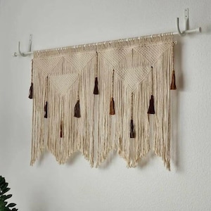 May include: A beige macrame wall hanging with a geometric design and hanging tassels. The wall decor is suspended from a white metal rod and features diamond-shaped patterns and varying shades of brown tassels.