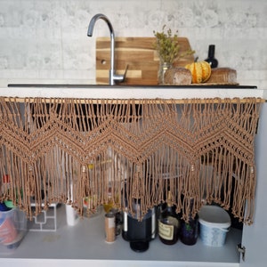 May include: A brown macrame curtain with fringe hangs over a kitchen cabinet. The curtain has a geometric pattern and is made of natural fibers. The cabinet is open and contains various items, including bottles, jars, and a coffee maker.