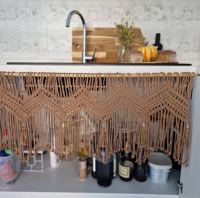 May include: A brown macrame curtain with fringe hangs over a kitchen cabinet. The curtain has a geometric pattern and is made of natural fibers. The cabinet is open and contains various items, including bottles, jars, and a coffee maker.