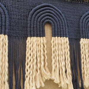 May include: A wall hanging with a woven design of two arches in navy blue with a fringe of cream colored yarn.