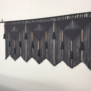 May include: Gray macrame wall hanging with a geometric pattern and wooden beads. The hanging has a fringe of tassels at the bottom.