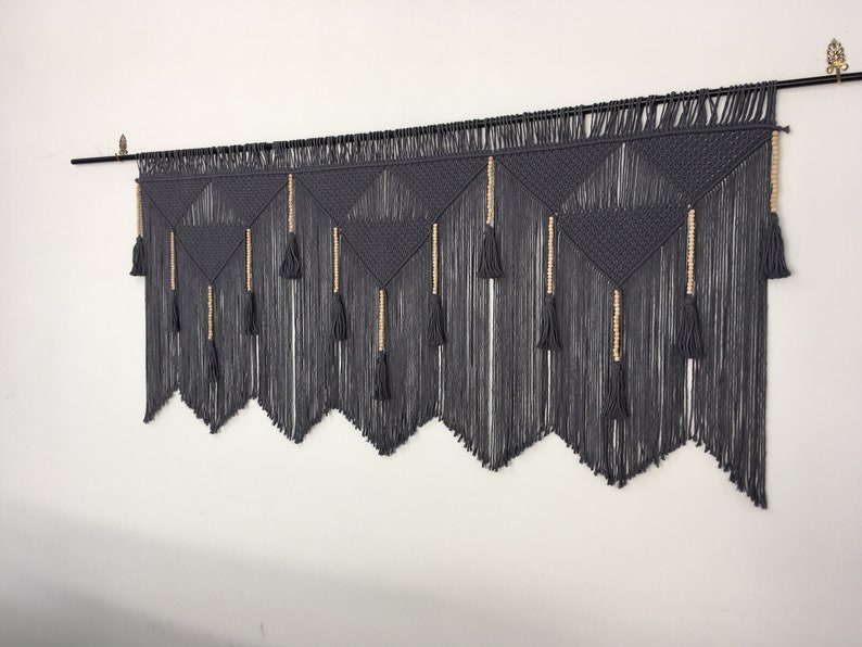 May include: Gray macrame wall hanging with a geometric pattern and wooden beads. The hanging has a fringe of tassels at the bottom.