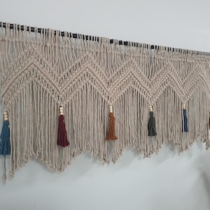 May include: A beige macrame wall hanging with a chevron pattern and five tassels in different colors: blue, burgundy, orange, green, and brown.