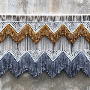 May include: A macrame wall hanging with a wooden dowel. The design features a repeating chevron pattern in white, mustard yellow, and gray. The bottom edge has a fringe detail. The wall hanging is displayed against a textured gray wall.