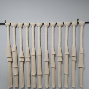 K&ouml;nnte beinhalten: Ein Wandbehang aus wei&szlig;em Makramee mit mehreren Garnstr&auml;ngen, die an einer schwarzen Stange h&auml;ngen. Die Str&auml;nge sind in einer Reihe von Knoten zusammengebunden, um einen dekorativen Quasten-Effekt zu erzeugen.