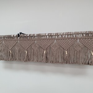 May include: A macrame valance in a neutral taupe color, featuring geometric diamond patterns and fringe. Wooden beads accent the design. The valance is suspended from a metal rod, adding a bohemian touch to home decor.