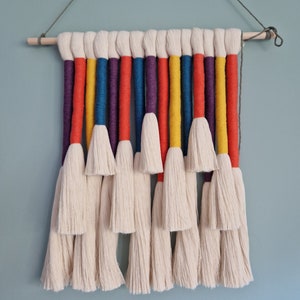 Può includere: Un arazzo da parete realizzato con filo bianco con strisce colorate in rosso, arancione, giallo, blu e viola. Il filo è legato insieme in una serie di nappe.
