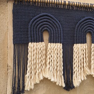 May include: A navy blue and cream macrame wall hanging with two arches. The hanging has long fringe on the bottom.