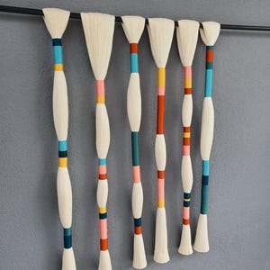 May include: A decorative wall hanging featuring seven off-white, brush-like elements suspended from a black rod. Each element has a colorful, cylindrical handle with bands of teal, pink, orange, and yellow. The background is a solid gray.