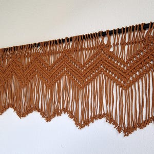 May include: A brown macrame wall hanging with a chevron pattern and fringe. The hanging is attached to a black rod.