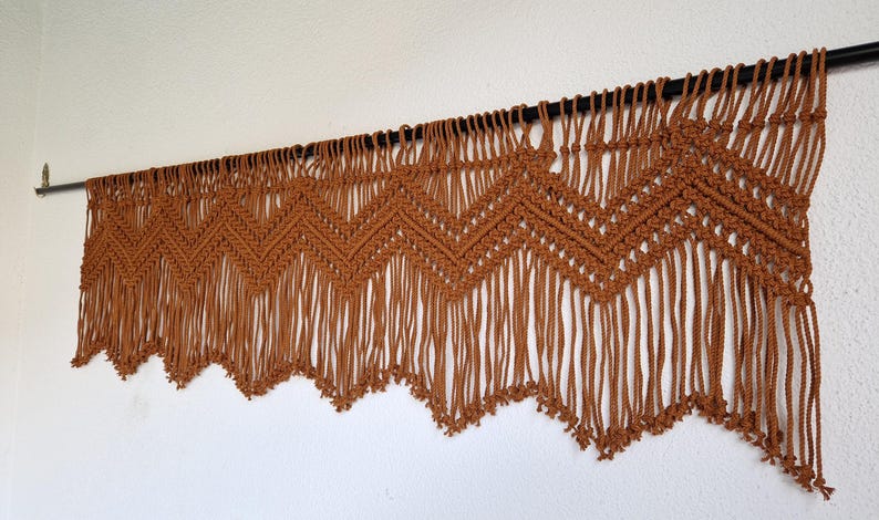 May include: A brown macrame wall hanging with a chevron pattern and fringe. The hanging is attached to a black rod.