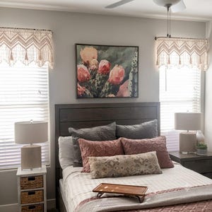 May include: A bedroom featuring a dark wood headboard, a bed with layered pillows in shades of gray and dusty rose, a wooden breakfast tray, and two windows adorned with cream-colored macrame valances. A framed cactus print hangs above the bed.