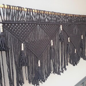 May include: A black macrame wall hanging with a wooden dowel. The design features geometric patterns, tassels, and white wooden beads. The macrame is made of thick, dark yarn and hangs against a white wall.