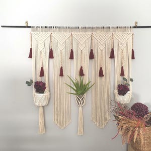 May include: A large cream-coloured macrame wall hanging suspended from a black rod, featuring intricate knotted patterns, wooden beads, and deep burgundy tassels. The piece includes three integrated plant holders containing green plants.