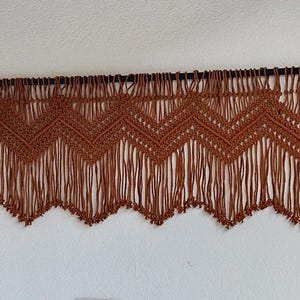 May include: A brown macrame wall hanging with a chevron pattern and fringe. The hanging is attached to a black rod with gold finials.