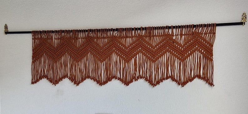 May include: A brown macrame wall hanging with a chevron pattern and fringe. The hanging is attached to a black rod with gold finials.