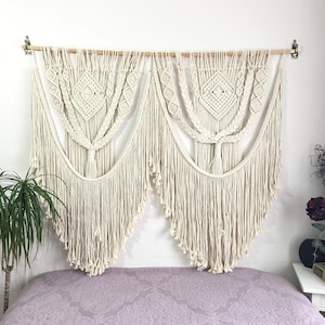 以下が含まれることがあります： 幾何学模様の大きな白いマクラメの壁掛け。壁掛けは綿のコードで作られており、上部に木製の棒があります。紫色のベッドスプレッドの上に吊り下げられています。