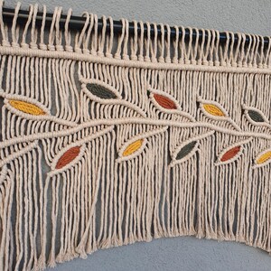May include: Macrame wall hanging featuring a branch design with colorful leaves. The leaves are in shades of yellow, orange, and green. The macrame is made of natural-colored cotton cord and hangs from a black rod.