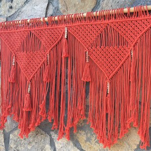 May include: A red macrame wall hanging with a geometric pattern and wooden beads. The hanging has a fringe of tassels.