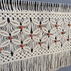 May include: A cream-colored macrame wall hanging featuring a repeating geometric knot pattern. The design incorporates small, round, speckled orange beads integrated into the center of each knot motif, finished with a long fringe at the bottom.