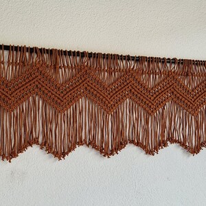 May include: A brown macrame wall hanging with a geometric pattern and fringe. The hanging is attached to a black rod.