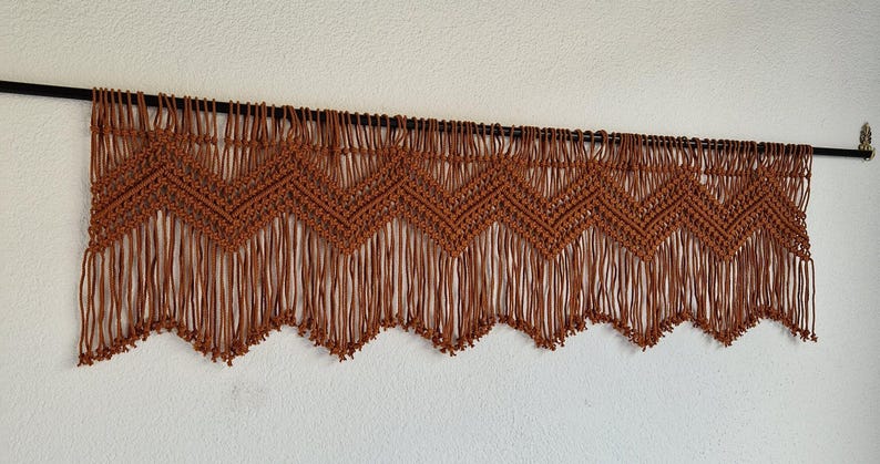 May include: A brown macrame wall hanging with a geometric pattern and fringe. The hanging is attached to a black rod.