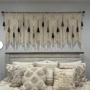 May include: A macrame wall hanging with a wooden rod, featuring a cream-colored woven design with triangular patterns and hanging tassels in black and gray. The bed below has decorative pillows with various textures and a pillow with the text "dogs are not our whole lives but they make our lives whole."