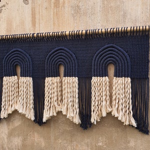 May include: A wall hanging made of woven macrame with three arches in a navy blue color. The arches are topped with a fringe of natural colored yarn.