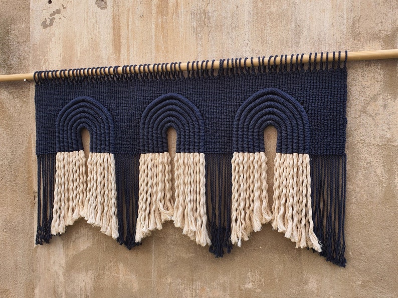May include: A wall hanging made of woven macrame with three arches in a navy blue color. The arches are topped with a fringe of natural colored yarn.