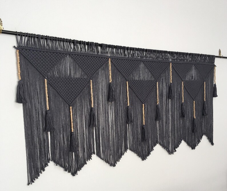 May include: A gray macrame wall hanging with wooden beads and tassels. The hanging is made of a thick, textured yarn and has a geometric pattern.